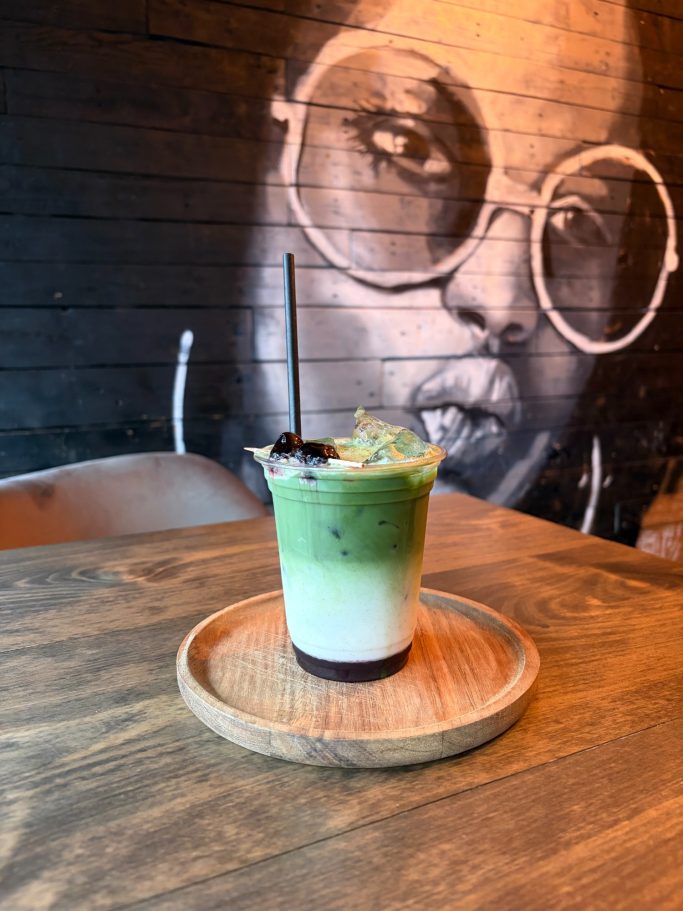 A layered drink in a glass on a wooden tray, with a mural of a woman in the background.