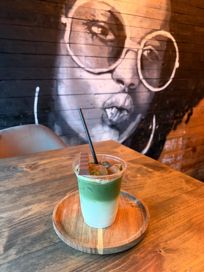 A layered green and white drink served on a wooden plate, with a mural of a woman in the background.